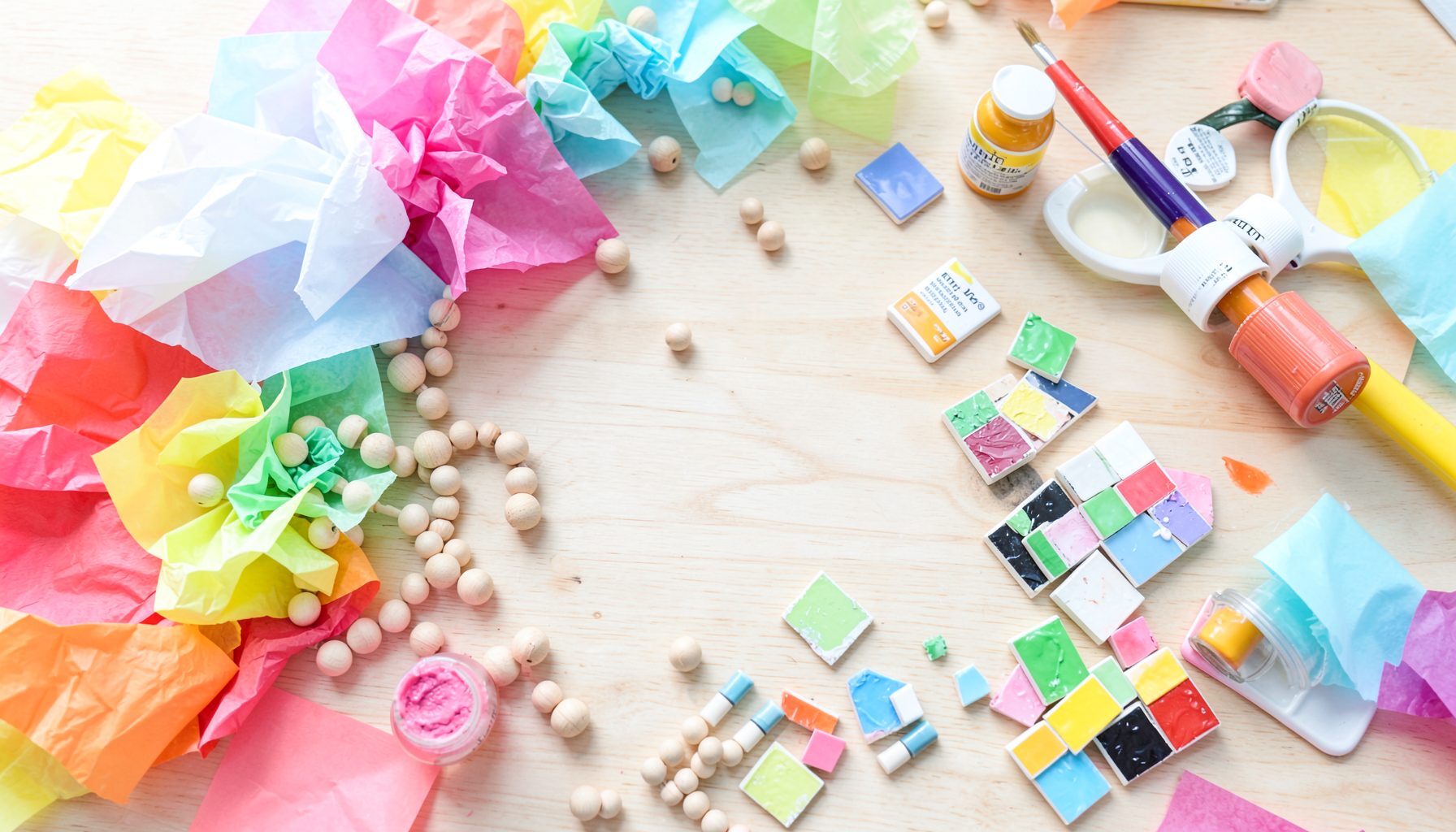 Overhead view of colorful tissue paper, wooden beads, small painted tiles, and a paintbrush arranged on a light wood surface